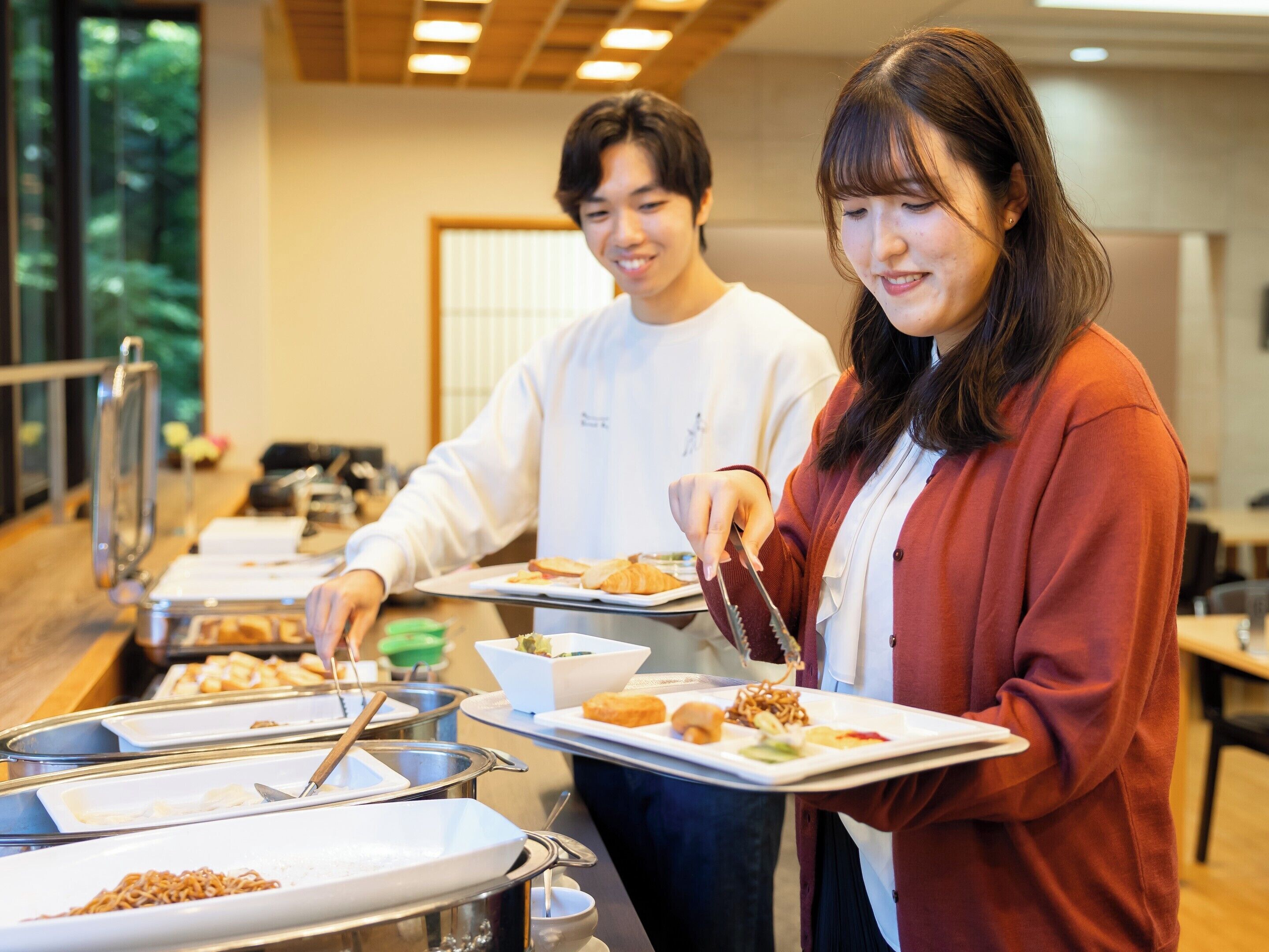 Disposición de un bufé con diferentes alimentos en recipientes metálicos, una persona sirviendo comida y otra sosteniendo una bandeja.