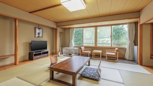 Ein traditioneller japanischer Raum mit Tatami-Bodenbelag, einem niedrigen Holztisch, einem Fernseher und einem großen Fenster mit Vorhängen.