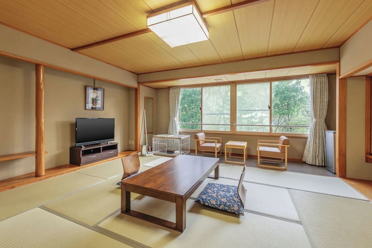 Une pièce de style japonais traditionnel, avec un sol recouvert de tatamis, une basse table en bois, un téléviseur et une grande fenêtre avec des rideaux.