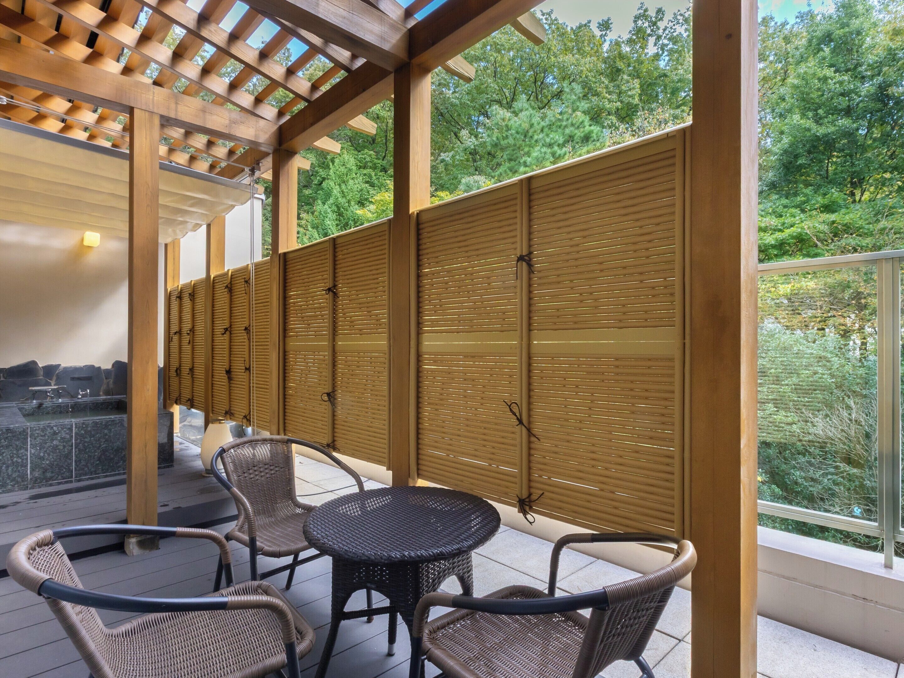 A covered outdoor seating area with wicker chairs and a round table, a stone water feature, and a view of a forested area.