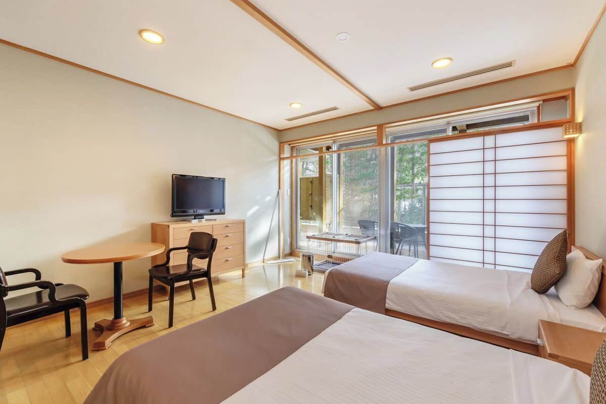 Une chambre d’hôtel avec deux lits, un bureau avec une chaise, une télévision et de grandes fenêtres équipées de stores.