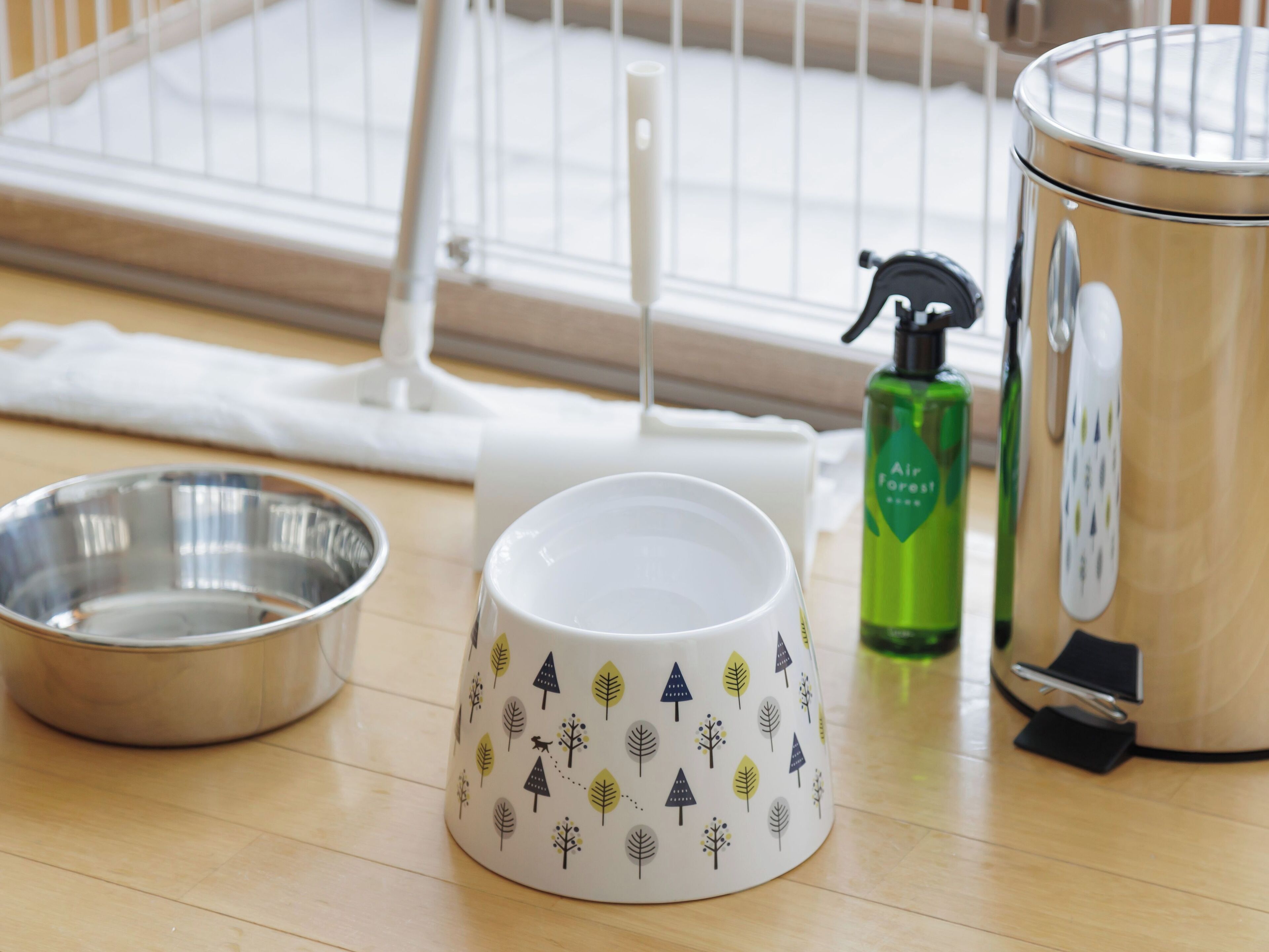 A pet bowl, a white mug with tree patterns, a green bottle, and a stainless steel container on a wooden floor.