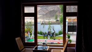 Una habitación con vistas a un paisaje montañoso a través de grandes puertas de cristal. Adentro, hay una silla de madera, una mesa pequeña con un vaso y un banco junto a la ventana.