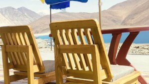 Dos sillones de madera bajo un paraguas de colores, con un paisaje montañoso al fondo.
