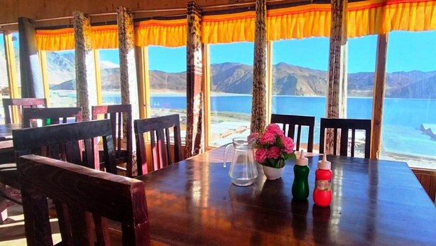 Una mesa de madera con sillas, una jarrón y botellas, frente a un paisaje con montañas y un lago.