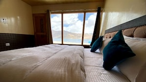 Habitación de hotel con una cama grande, vistas a montañas y un lago a través de una ventana, y una puerta con la leyenda 'SKY MAGNIFICENT'.