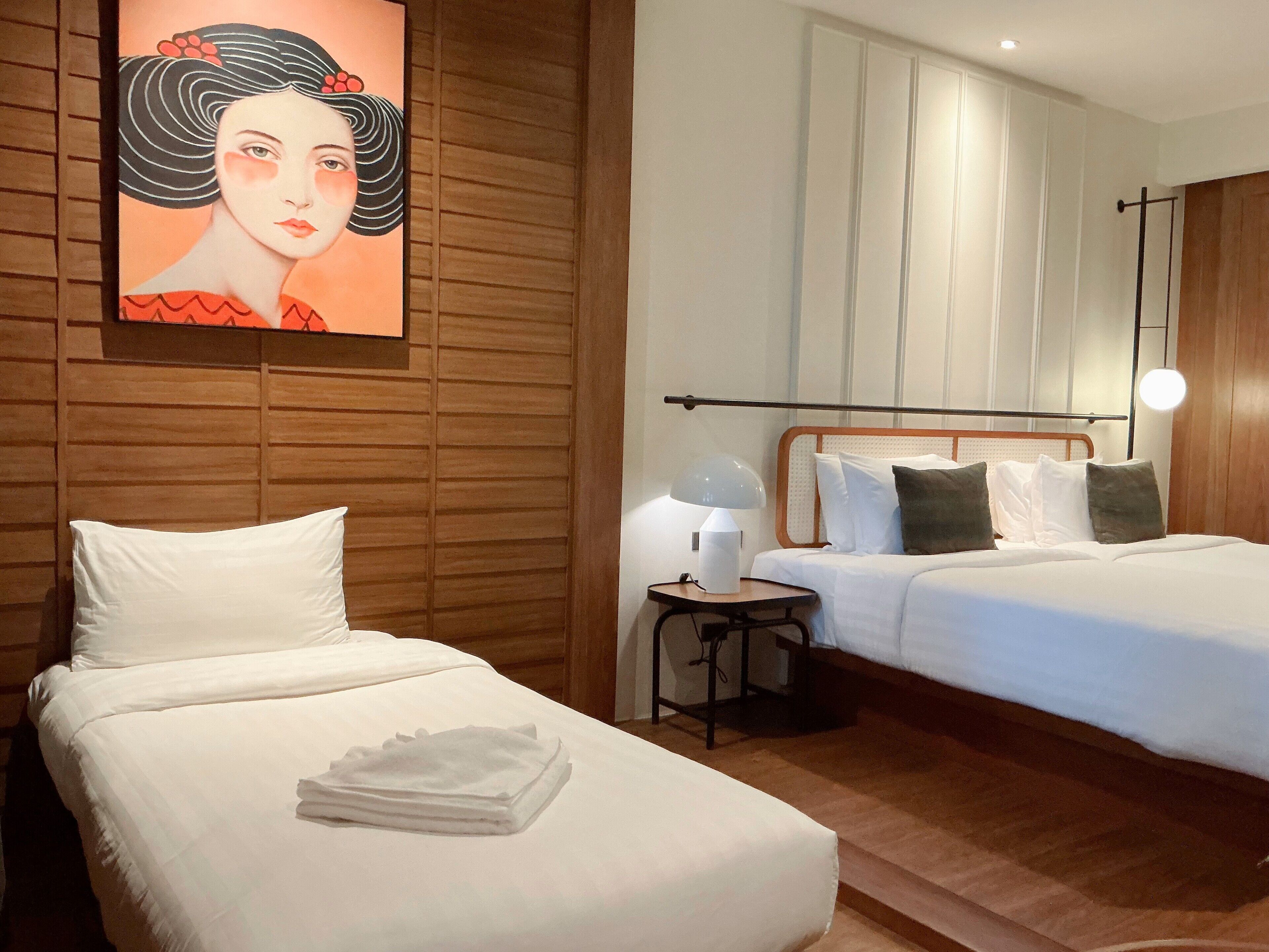 A hotel room with two beds, a painting on the wall, a bedside table with a lamp, and a wooden headboard.