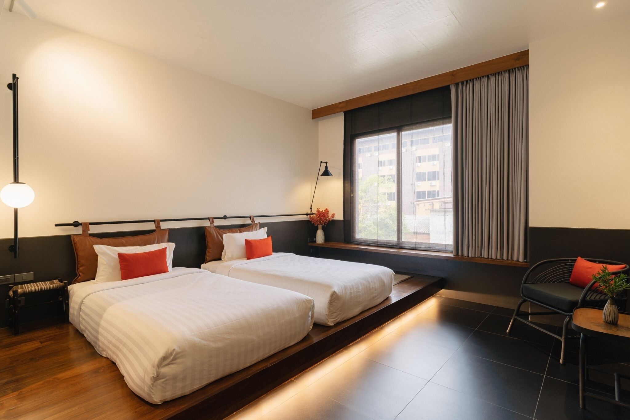 A hotel room with two beds, a sitting area with a chair, a small table, a lamp, and a window with curtains.