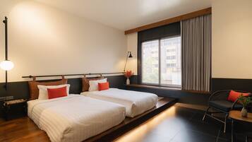 A hotel room with two beds, a sitting area with a chair, a small table, a lamp, and a window with curtains.