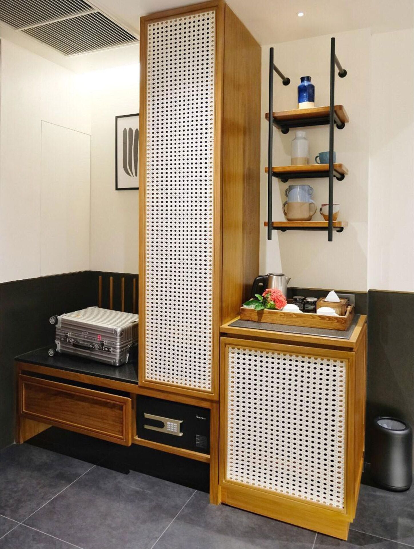 Een kamer met een hoge, geperforeerde houten kast, een klein plankje met bekers, een koffer en een prullenbak.