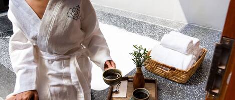 Una persona vestida con una bata blanca sentada en un banco de spa, con una bandeja que contiene una taza, un cuenco y una pequeña planta. Hay toallas apiladas en una cesta y se aprecia el marco de una puerta de madera.