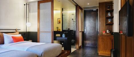 A modern hotel room with a large bed, a wooden wardrobe, a bathroom with a shower, and a small kitchenette.