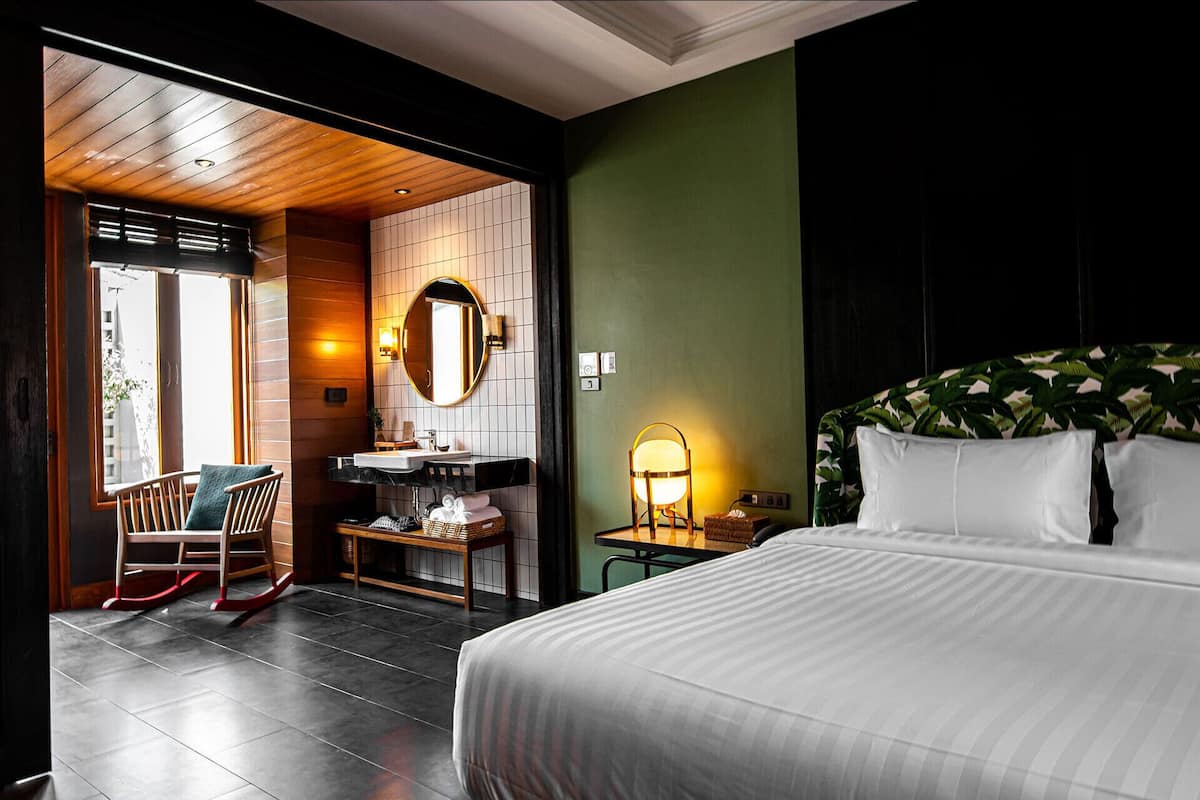 Une chambre d’hôtel avec un lit, un fauteuil à bascule en bois, une salle de bain avec un lavabo et un miroir, et une vue sur l’extérieur.