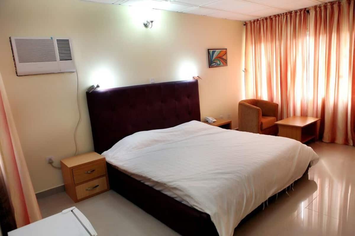 A hotel room with a bed, a chair, a bedside table, a nightstand, an air conditioning unit, and a painting on the wall.