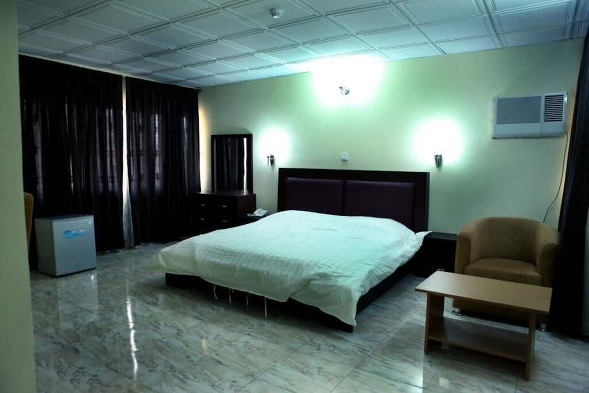 A hotel room with a large bed, a bedside table, a chair, a wardrobe, and an air conditioning unit.