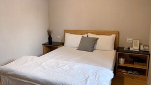 Een netjes opgemaakt bed met witte beddengoed en een grijs kussen, een nachtkastje met een telefoon en een lamp, en een klein plankje met slippers.