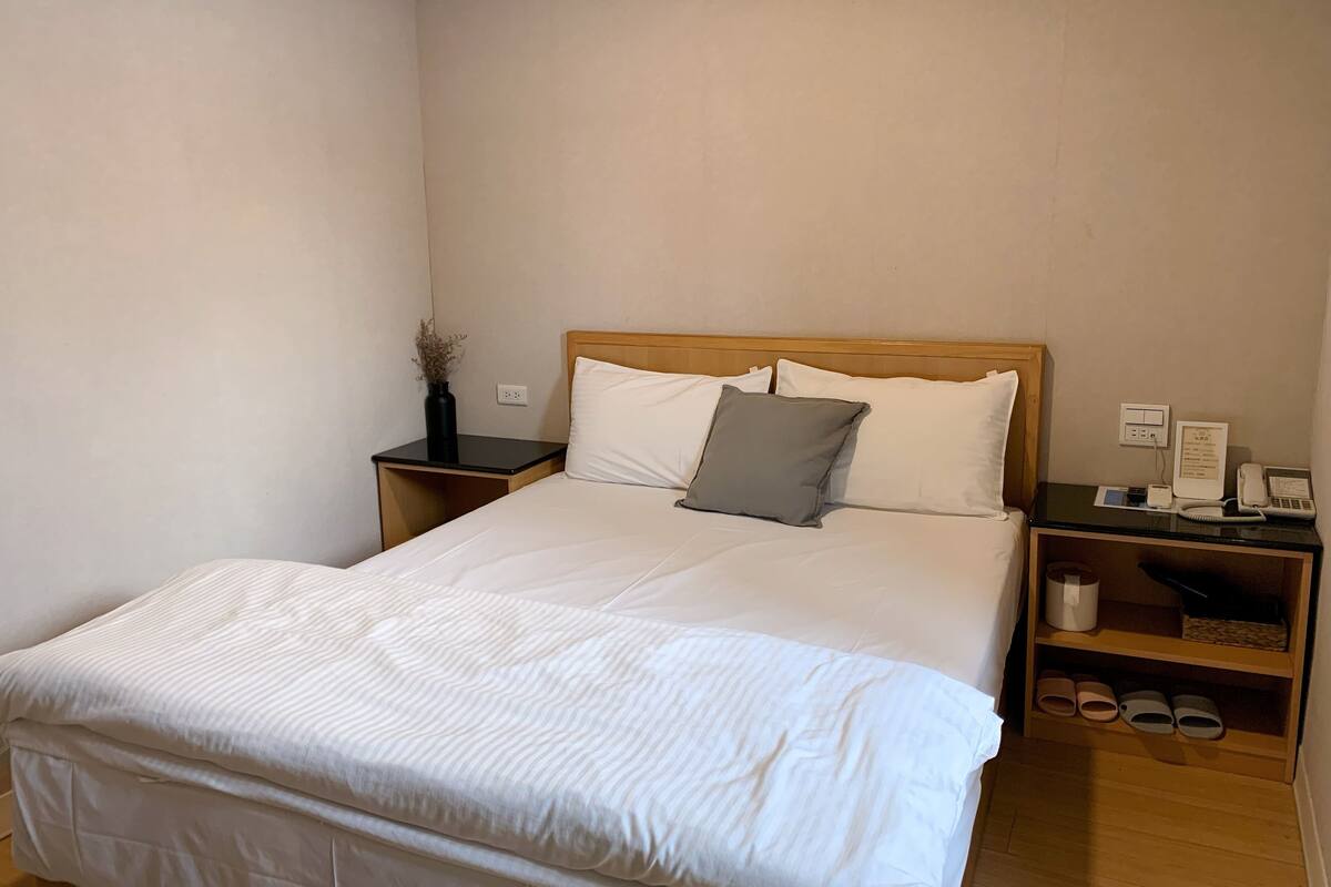 Ein ordentlich bezogenes Bett mit weißer Bettwäsche und einem grauen Kopfkissen, ein Nachttisch mit Telefon und Lampe sowie ein kleines Regal mit Hausschuhen.