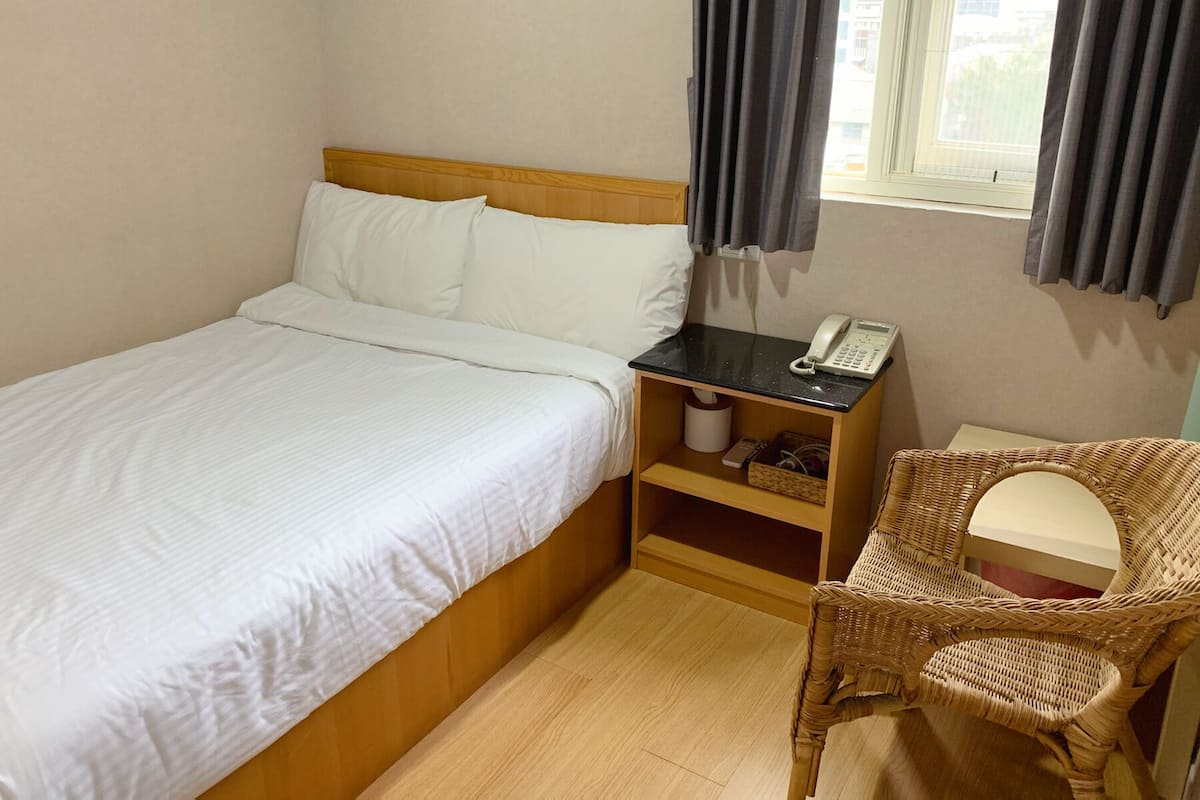 Ein Hotelzimmer mit einem Bett, einem Nachttisch mit Telefon, einem Korbstuhl und einem Vorhangfenster.