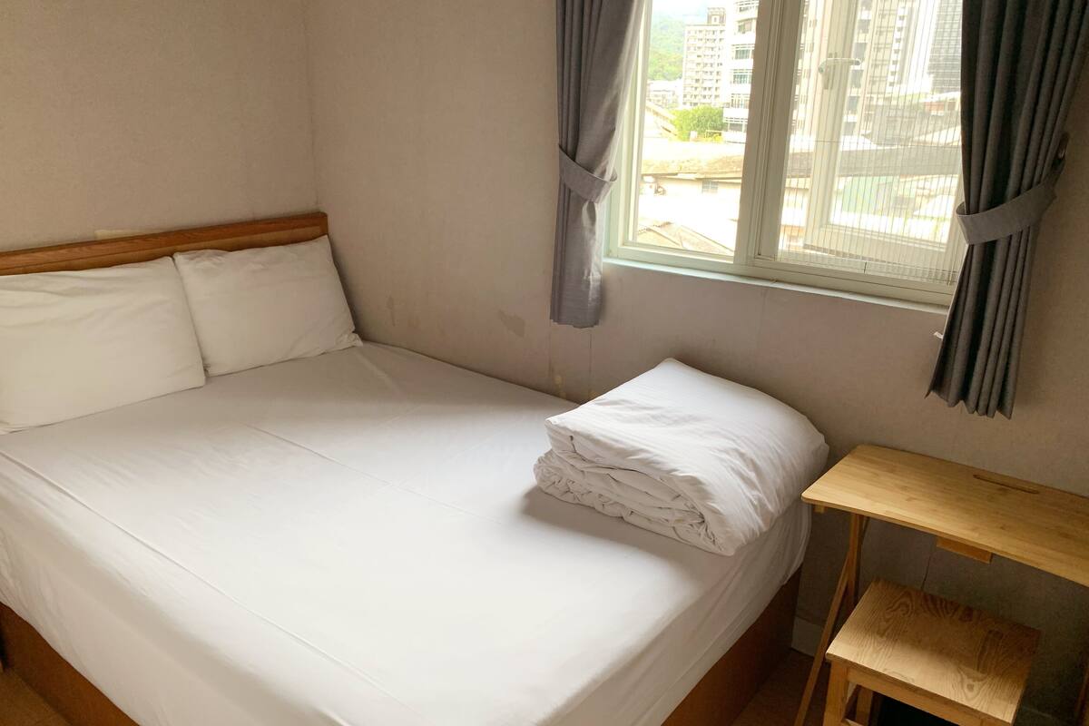 Ein Zimmer mit einem Einzelbett, einem Holzkopfstück, einem kleinen Holztisch und einem Fenster mit Vorhängen.