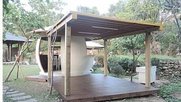 Une structure de terrasse en bois surmontée d’une pergola, entourée d’un jardin avec un mur de pierres et des arbres.
