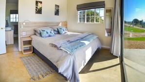 A bedroom with a bed, bedside tables, a window with a view, and an air conditioning unit. - Addo Park Vista Cottages (Colchester)