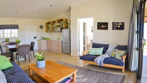 A spacious living room with a sofa, a coffee table, and a dining area with a table and chairs. There is a kitchen area with cabinets and a refrigerator. - Addo Park Vista Cottages (Colchester)