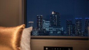 A hotel room with a cityscape view through a window, a bed with pillows, and a control panel on the wall. - Hotel Vista Tokyo Tsukiji (Tokyo)
