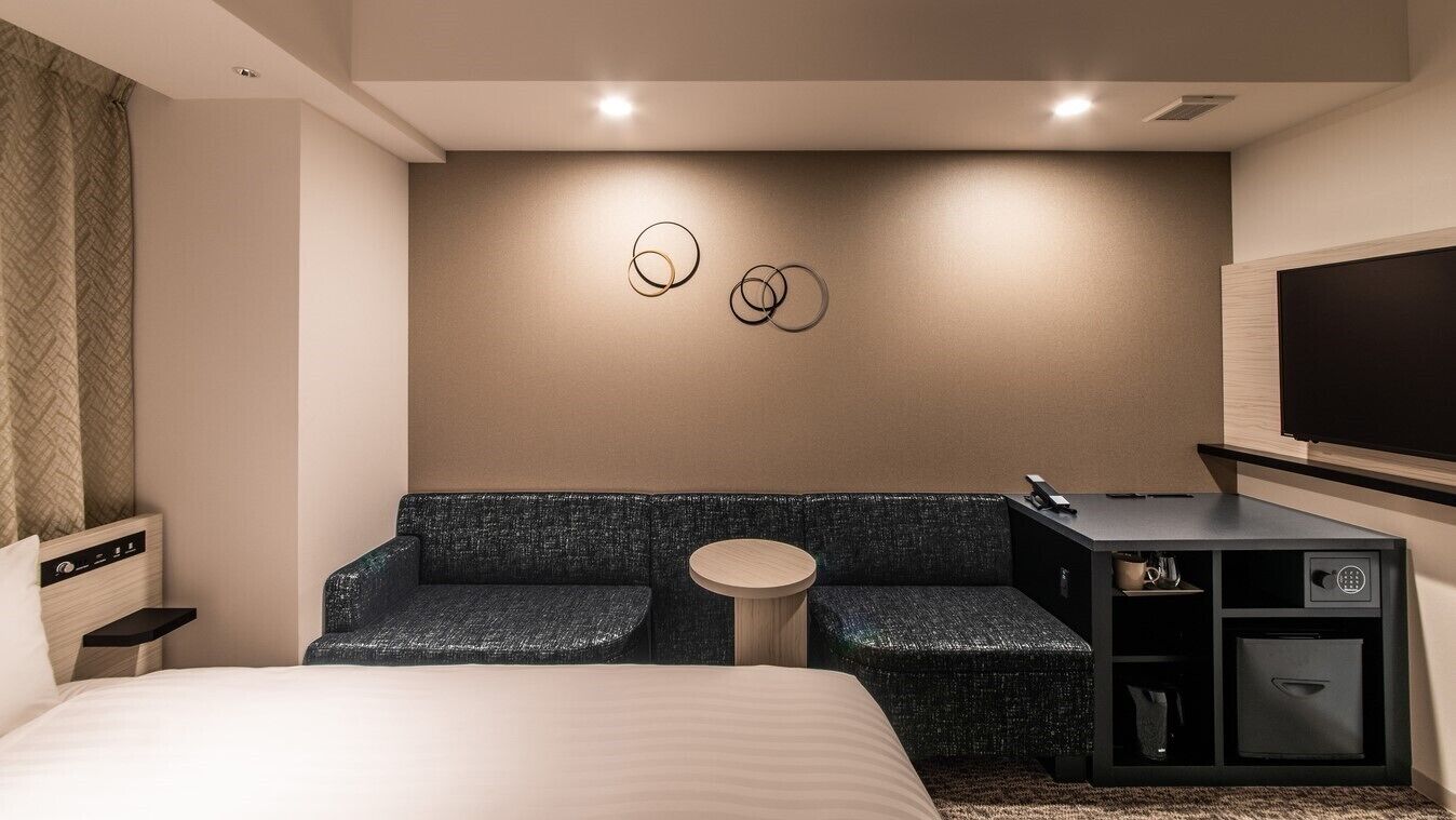 A modern hotel room with a bed, a seating area with a round table, a TV, and a minibar.