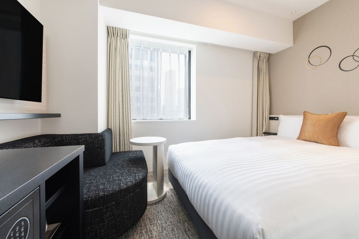 Habitación de hotel con cama, televisor, zona de estar y ventana con cortinas.