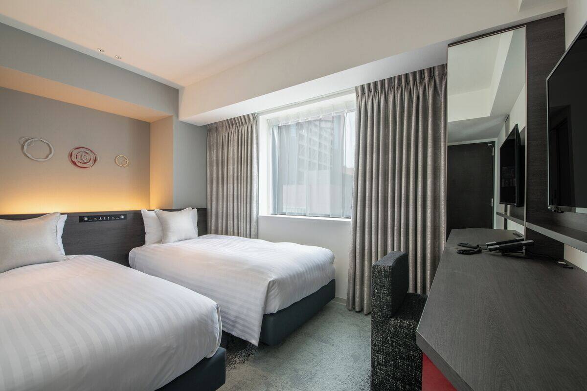 Habitación de hotel con dos camas, un escritorio con televisor de pantalla plana, un ventanal con cortinas y un espejo montado en la pared.