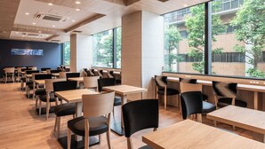 A modern dining area with wooden tables and chairs, large windows with a view of a brick building and trees, and a blue wall with a framed picture. - Hotel Vista Tokyo Tsukiji (Tokyo)