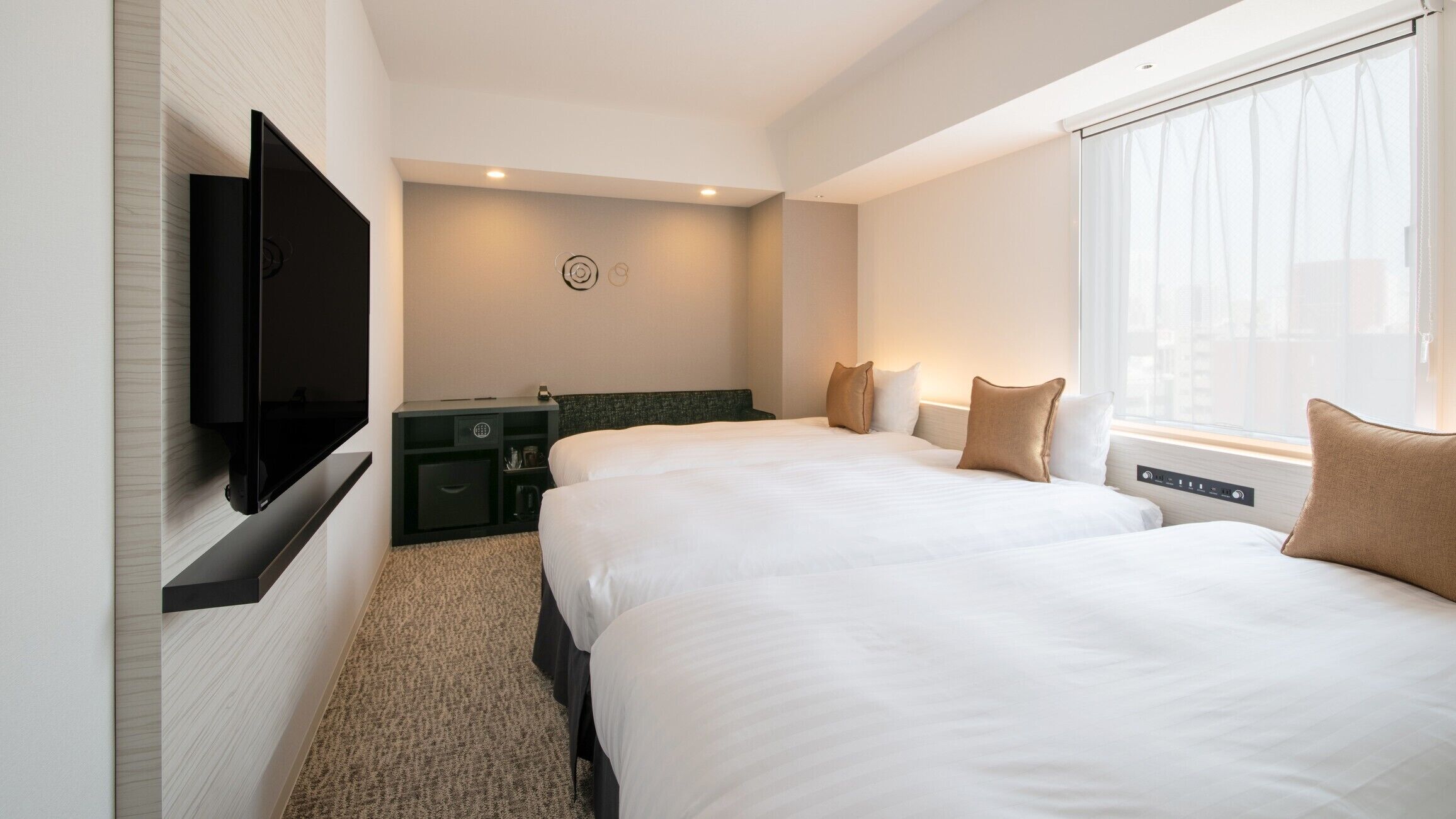 A hotel room with two beds, a flat-screen TV mounted on the wall, a small desk with a lamp, and a window with curtains.