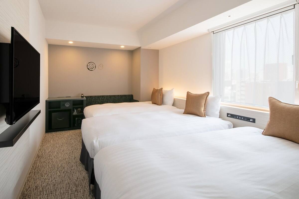 Habitación de hotel con dos camas, un televisor de pantalla plana montado en la pared, un escritorio pequeño con una lámpara y una ventana con cortinas.