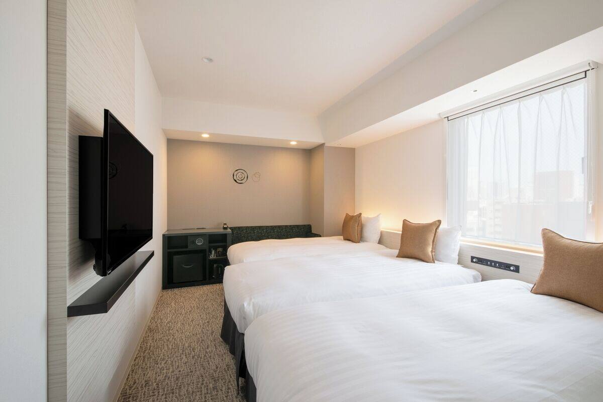 Habitación de hotel con dos camas, un televisor de pantalla plana montado en la pared, un escritorio pequeño y una ventana con cortinas.