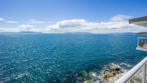 Une vue dégagée sur la mer depuis un balcon doté d’une balustrade, avec une côte rocheuse et des montagnes lointaines sous un ciel bleu.