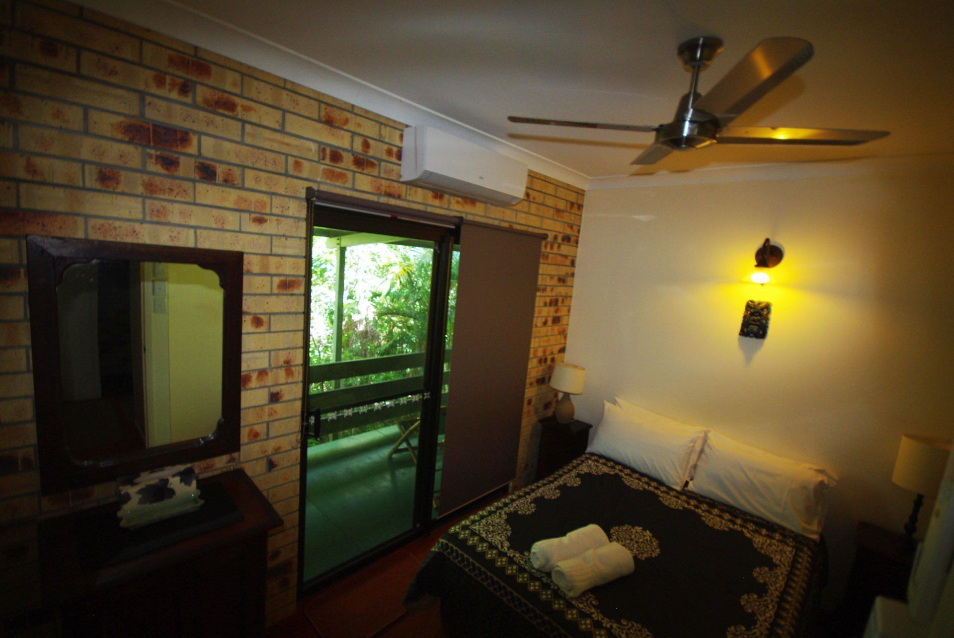 Une chambre d’hôtel avec un mur en briques, un ventilateur de plafond, un lit recouvert d’une courtepointe à motifs, une table de chevet avec une lampe et une vue sur la verdure par une porte.
