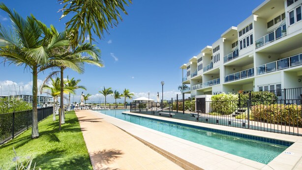 A swimming pool with a tiled border, surrounded by a fence, and adjacent to a multi-story building with balconies. - Beachside Apartment 5 (Nelly Bay)