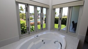A modern bathroom with a large bathtub, large windows, and a view of a pool and garden. - Beachside Apartment 5 (Nelly Bay)