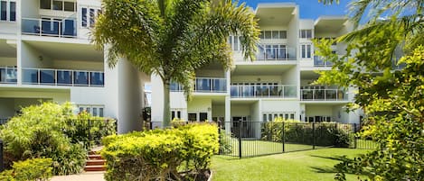 A multi-story residential building with balconies, surrounded by well-maintained greenery and a clear sky.