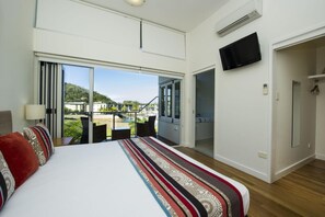 A bedroom with a large bed, a TV mounted on the wall, and a balcony with a view of a pool and buildings.