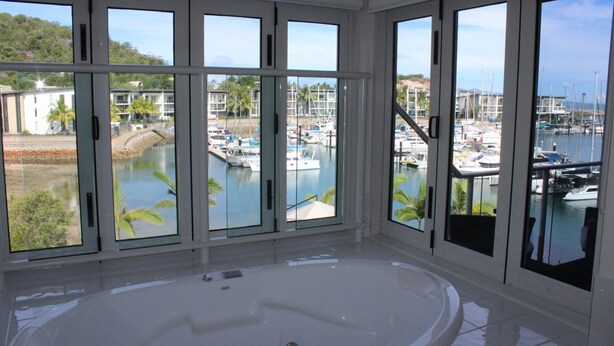 Een ruime badkamer met een groot bad en meerdere ramen met uitzicht op een jachthaven en omliggende gebouwen.