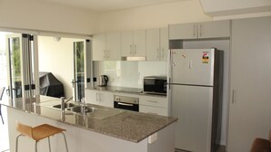 Een moderne keuken met witte kasten, een roestvrijstalen koelkast, een magnetron en een koffiezetapparaat. Er is een barstoel en een raam met uitzicht op groen.