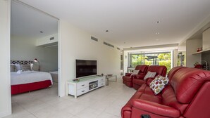 A spacious living room with a red leather sofa, a flat-screen TV, and a dining area. - Blue on Blue Apartment 1015 (Nelly Bay)