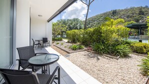 A modern outdoor patio with a glass table and chairs, surrounded by a gravel garden and a view of a hillside. - Blue on Blue Apartment 1015 (Nelly Bay)