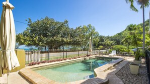 A rectangular swimming pool with a brick edge, surrounded by a pebble path and a low brick wall. There are outdoor chairs and a table near the pool, and a palm tree in the background. - Champagne Apartment 1 (Arcadia)