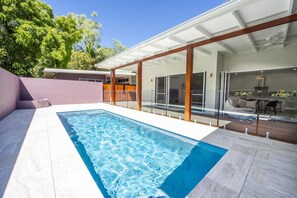 Una piscina moderna al aire libre con un elemento acuático de agua azul cristalina, rodeada por una terraza de madera y un área de patio con cerramiento de vidrio.