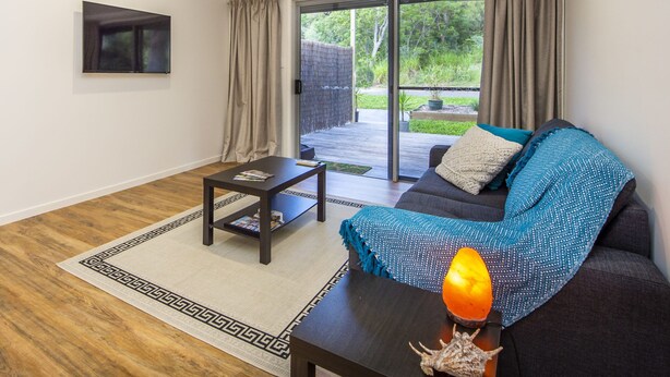 A modern living room with a dark sofa, a coffee table, and a view of a garden through a sliding glass door. - Sail Rock Apartment 1 (Picnic Bay)