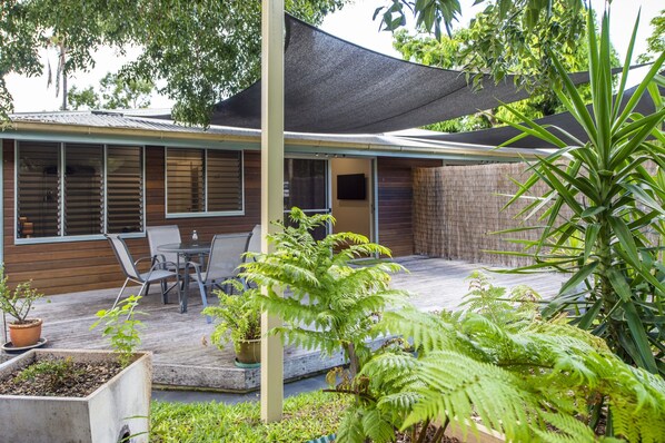 A covered patio with a table and chairs, surrounded by various plants and a large tree.