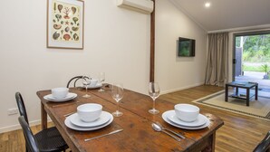 Une salle à manger avec une table en bois dressée pour quatre personnes, des assiettes blanches et des verres à vin. Un cadre représentant une illustration botanique est accroché au mur, ainsi qu’un appareil de climatisation. On peut également admirer la vue sur l’extérieur à travers une porte vitrée.