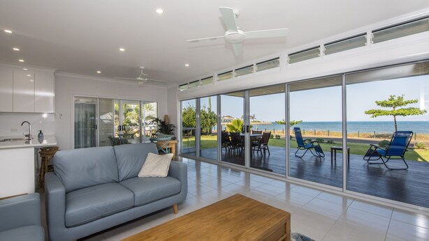 Una sala de estar moderna con un sofá gris, una mesa de centro de madera y grandes puertas correderas de vidrio que dan a una terraza con asientos exteriores y vista al mar.
